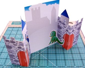 Puede incluir: Una tarjeta de castillo emergente con un panel central y dos torres laterales. La tarjeta presenta un cielo azul, siluetas de castillo y una pegatina de dragón verde. Las torres tienen puertas rojas y son de piedra gris.