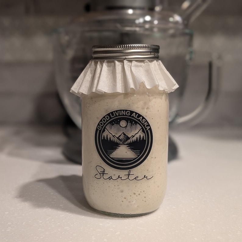 May include: A glass jar with a white cloth lid and a black and white label that says "Good Living Alaska Starter". The label features a mountain range and a lake.