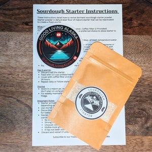 May include: A small orange pouch with a white label that says "Sourdough Starter" and has a logo of a mountain range with a red sun and a blue lake. The pouch is sitting on a white sheet of paper with instructions for reviving sourdough starter powder. The instructions include the text "Good Living Alaska" and "@GoodLivingAlaska".