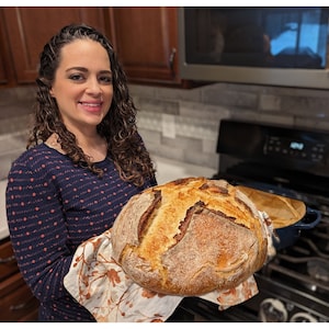 May include: A woman holds a freshly baked loaf of sourdough bread in a kitchen setting. The bread has a golden brown crust and a soft, airy interior.