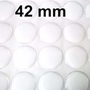 Puede incluir: Una colección de cabujones redondos de vidrio transparente, cada uno de aproximadamente 4,2 cm de diámetro, dispuestos sobre una superficie blanca. El texto "42 mm" se muestra en negro, indicando el tamaño de los cabujones.
