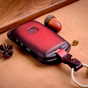 Puede incluir: Una funda de llavero de cuero burdeos con un llavero dorado y una etiqueta de cuero a juego que dice "Beth". La funda tiene un tono rojo oscuro y está junto a una bellota y una flor seca.