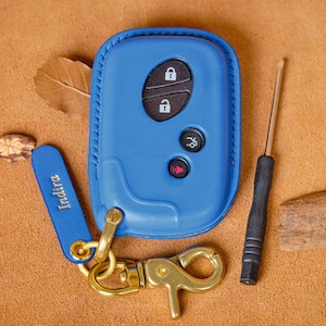 May include: A blue leather key fob cover with a gold-colored clasp and a matching blue tag that reads "Indira". The cover has black buttons and a key fob. A small screwdriver and wood pieces are also present.