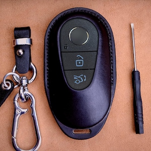 Puede incluir: Una funda de cuero negro para la llave del coche con un llavero y un pequeño destornillador. El llavero tiene tres botones y un ojo de cerradura. Una etiqueta de cuero negro con el nombre "Stiles" está unida al llavero.