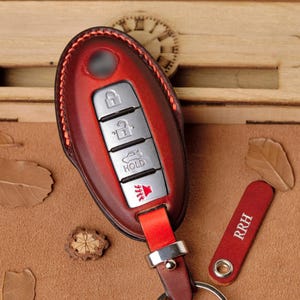 May include: A red leather car key fob cover with a silver key ring. The fob has silver buttons and a red leather strap. A matching red leather tag with "RRH" is also included. The key fob is on a brown surface.