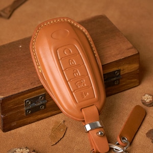 May include: A tan leather car key fob cover with embossed buttons and a key ring. The cover is on a brown surface with a wooden box and decorative elements.