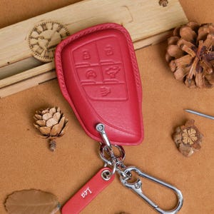 May include: A red leather car key fob cover with a silver keyring and a red tag that says "Leo". The fob cover has embossed buttons and is surrounded by decorative items on a brown surface.