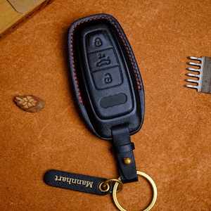 May include: Black leather car key fob cover with red stitching, featuring key buttons. Includes a gold-colored keyring and a black tag with the name "Mannhart". The fob is on a brown leather surface.