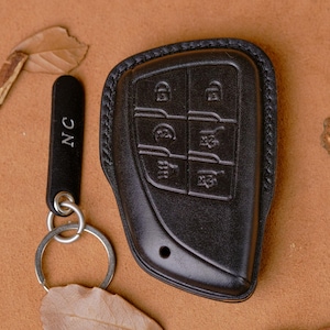 May include: Black leather car key fob cover with a matching keychain. The key fob cover has embossed buttons and a stitched border. The keychain has the initials "NC" and a silver key ring.