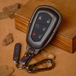 May include: A dark brown leather key fob cover with white stitching, featuring buttons for lock, unlock, and other car functions. Includes a small tag with text and a metal clasp. The fob is on a brown surface.