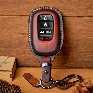 May include: A brown leather key fob cover with a key ring and a small leather tag. The key fob has black buttons with white icons and the word "HOLD". The cover is stitched with a lighter brown thread.