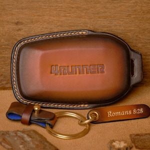 Puede incluir: Un juego de llavero y llavero de cuero marrón. El llavero está grabado con la palabra "4RUNNER". La etiqueta del llavero dice "Romanos 8:28". El juego tiene un llavero dorado y un interior azul.