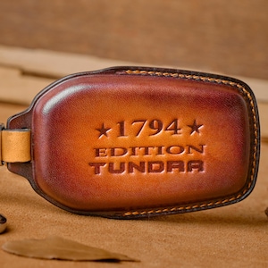 May include: A brown leather key fob with the text "1794 EDITION TUNDRA" embossed on the front. The fob has a metal keyring and a leather strap. The leather has a gradient colour from light to dark brown.