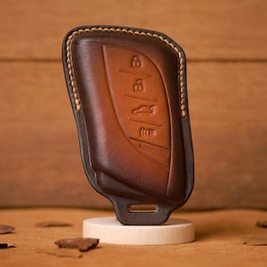 Puede incluir: Una funda de cuero marrón para la llave del coche con un borde cosido e iconos en relieve para bloquear, desbloquear, maletero y otras funciones. La funda se muestra sobre un pequeño soporte blanco sobre un fondo marrón.