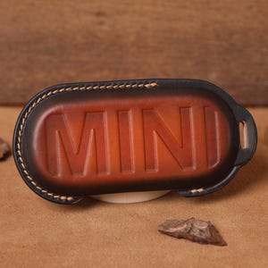 May include: A brown leather key fob with the embossed word "MINI". The key fob has a black border with beige stitching. The leather has a gradient effect, transitioning from dark brown to a lighter brown in the center.