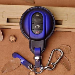 Può includere: Una custodia per portachiavi per auto in pelle blu con moschettone argentato e cinturino in pelle blu abbinato. Il portachiavi presenta icone in rilievo e una combinazione di colori blu e nero. Il portachiavi è mostrato su una superficie marrone con foglie.