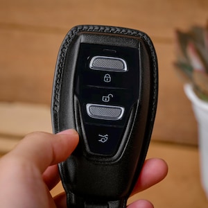 May include: Black leather car key fob cover with silver-colored buttons. The cover has a stitched border and features lock, unlock, and trunk release symbols. The key fob is held in a person's hand.