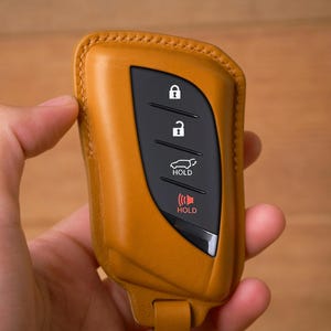 May include: A tan leather car key fob cover with a black control panel. The panel features lock, unlock, car, and hold buttons. The key fob is held in a hand against a blurred wooden background.