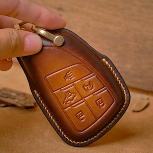 May include: A brown leather key fob cover with embossed icons for keyless entry. The cover has a gold-colored metal clasp and a stitched border. The leather has a gradient color from light to dark brown.