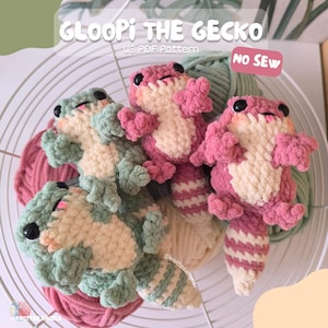 Puede incluir: Peluche de gecko tejido a ganchillo en tonos rosa, verde y crema, con ojos negros y colas a rayas. El texto "GLOOPI THE GECKO" y "US PDF Pattern" son visibles. Los peluches están dispuestos con hilo en una cesta de alambre.