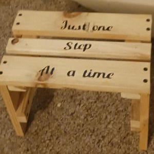 Puede incluir: Un pequeño taburete de madera clara con las palabras "Just one Step At a time" en negro. El taburete tiene un diseño sencillo y funcional, con tres escalones.