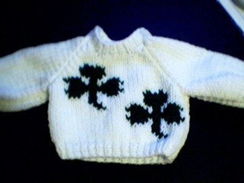 May include: White knitted baby sweater with black and green shamrock designs.
