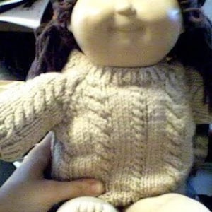 May include: A knitted beige sweater with a cable knit pattern. The sweater is worn by a doll with brown hair.