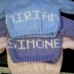 May include: Three knitted baby sweaters in different colors: light blue, purple, and pink. Each sweater has a white knitted name on the front: "TRIFECTA", "SIMONE", and "MIA".