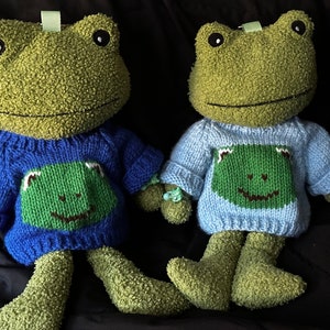 Puede incluir: Dos juguetes de peluche de rana verde que llevan suéteres de punto. Una rana lleva un suéter azul con un diseño de rana verde, y la otra lleva un suéter azul claro con un diseño de rana verde.