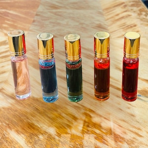 May include: Five glass roll-on bottles with gold caps, each filled with a different colored liquid: pink, blue, green, orange, and red. The bottles are arranged on a light brown wooden surface.