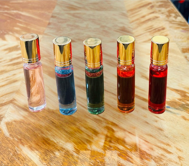 May include: Five glass roll-on bottles with gold caps, each filled with a different colored liquid: pink, blue, green, orange, and red. The bottles are arranged on a light brown wooden surface.