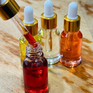May include: A close-up of several small glass bottles filled with colorful liquids. One bottle is open, with a dropper dispensing a red liquid. The other bottles contain yellow, clear, and orange liquids. Each bottle has a gold cap and a white dropper.