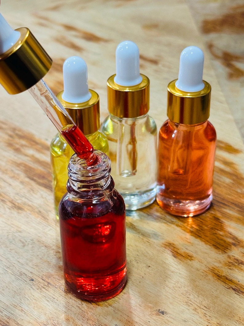 May include: A close-up of several small glass bottles filled with colorful liquids. One bottle is open, with a dropper dispensing a red liquid. The other bottles contain yellow, clear, and orange liquids. Each bottle has a gold cap and a white dropper.