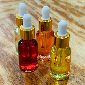 May include: Four small glass dropper bottles filled with colorful liquids. The bottles have gold-colored caps and white droppers. The liquids are various shades of red, orange, and yellow. The bottles are arranged on a wooden surface.