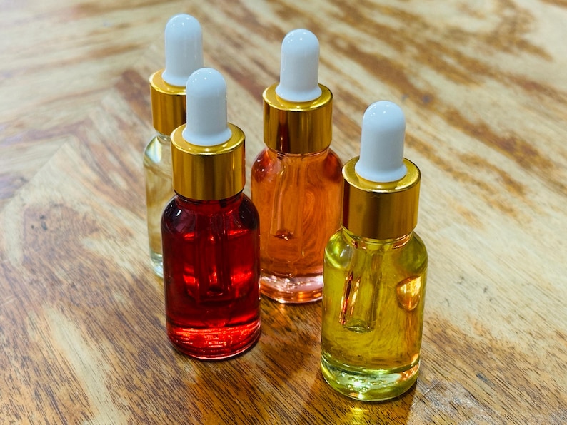 May include: Four small glass dropper bottles filled with colorful liquids. The bottles have gold-colored caps and white droppers. The liquids are various shades of red, orange, and yellow. The bottles are arranged on a wooden surface.