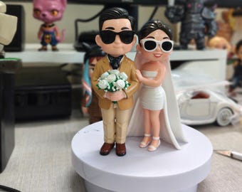 May include: A wedding cake topper featuring a bride and groom figurine. The groom wears a tan suit and sunglasses, holding a bouquet. The bride wears a white dress and veil, also with sunglasses. The figures stand on a white base.
