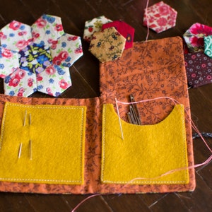May include: A small orange fabric pouch with two yellow felt pockets, one with needles and the other with sewing pins. The pouch is decorated with floral fabric and is surrounded by colorful fabric scraps.