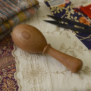 May include: A wooden knitting needle knocker with a smooth, rounded head and a long, slender handle. The knocker is made of light brown wood and has a natural finish.