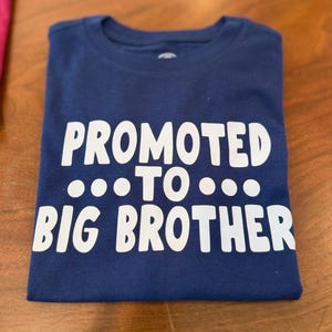 May include: A navy blue t-shirt with the words "PROMOTED TO BIG BROTHER" printed in white. The shirt is folded neatly and ready to be worn. A perfect gift for a new big brother.