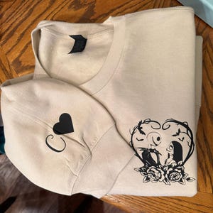 Pu&ograve; includere: Felpa beige con un cuore nero e la lettera "C" sulla manica. La parte anteriore presenta una grafica nera di Jack e Sally di The Nightmare Before Christmas all'interno di una cornice a forma di cuore con rose.