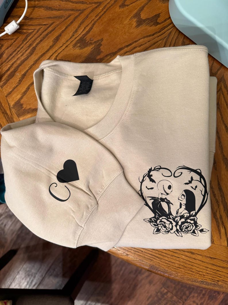 Pu&ograve; includere: Felpa beige con un cuore nero e la lettera "C" sulla manica. La parte anteriore presenta una grafica nera di Jack e Sally di The Nightmare Before Christmas all'interno di una cornice a forma di cuore con rose.