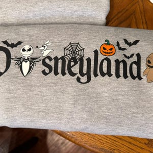 May include: A gray sweatshirt featuring the word "Disneyland" in a stylized font, with Halloween-themed graphics. The design includes bats, a spiderweb, a jack-o'-lantern, and characters like Jack Skellington and Oogie Boogie.