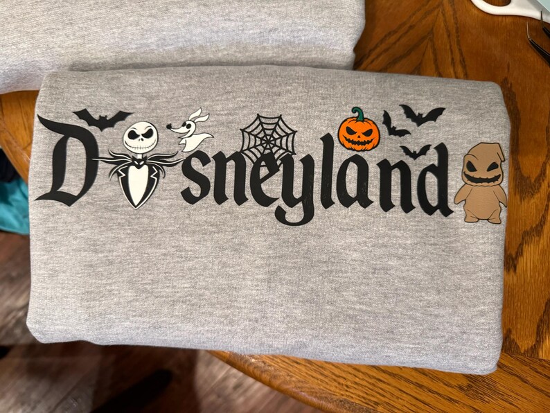 May include: A gray sweatshirt featuring the word "Disneyland" in a stylized font, with Halloween-themed graphics. The design includes bats, a spiderweb, a jack-o'-lantern, and characters like Jack Skellington and Oogie Boogie.