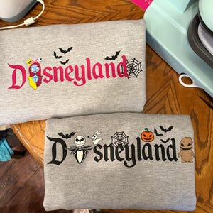 May include: Two gray sweatshirts with the word "Disneyland" in a stylized font. One features Sally from The Nightmare Before Christmas, bats, and a spiderweb. The other has Jack Skellington, Zero, a pumpkin, bats, a spiderweb, and Oogie Boogie.