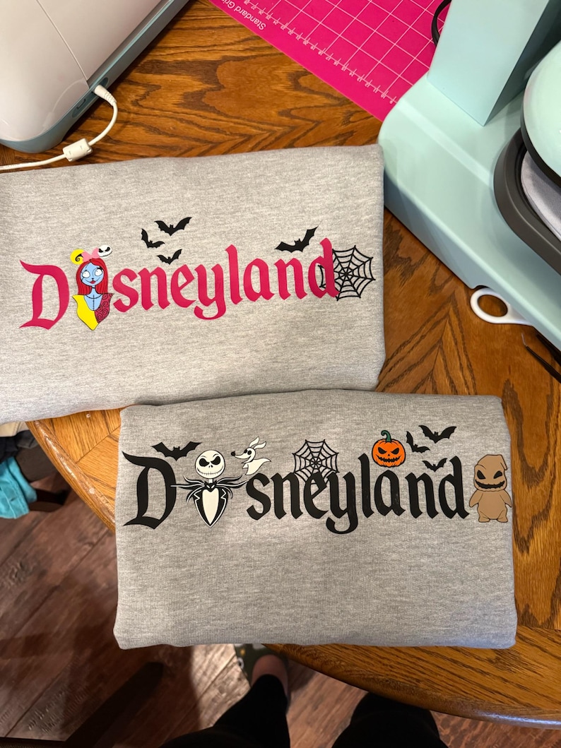 May include: Two gray sweatshirts with the word "Disneyland" in a stylized font. One features Sally from The Nightmare Before Christmas, bats, and a spiderweb. The other has Jack Skellington, Zero, a pumpkin, bats, a spiderweb, and Oogie Boogie.