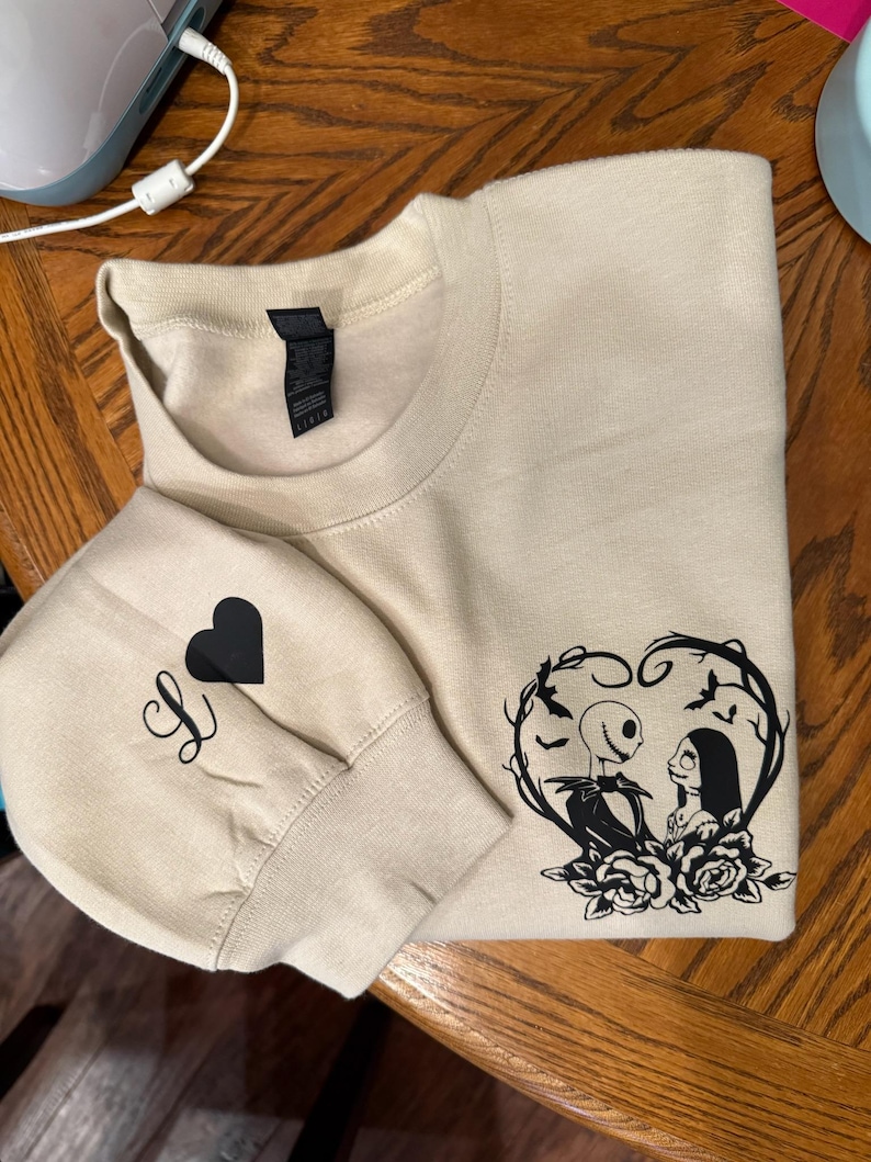 Pu&ograve; includere: Felpa beige con un cuore nero e la lettera "S" sulla manica. La parte anteriore presenta una grafica nera di Jack e Sally di The Nightmare Before Christmas all'interno di un design a forma di cuore con rose.