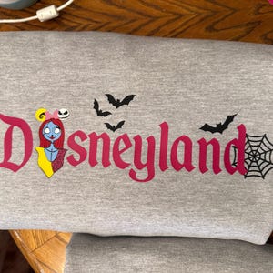 May include: A gray sweatshirt with the word "Disneyland" in pink, featuring a Sally character graphic, bats, and a spiderweb. The design has a Halloween theme.