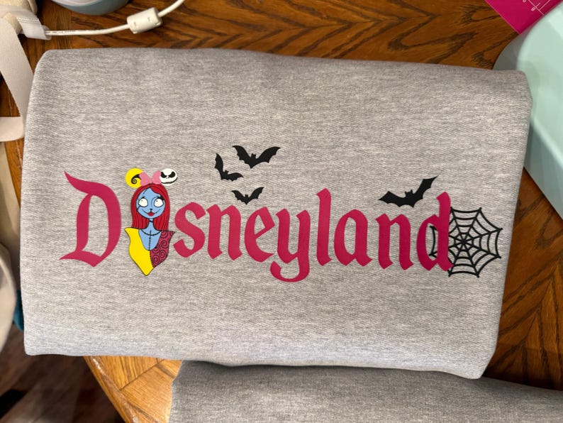 May include: A gray sweatshirt with the word "Disneyland" in pink, featuring a Sally character graphic, bats, and a spiderweb. The design has a Halloween theme.