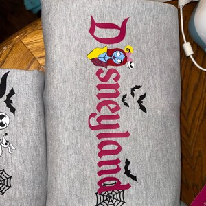 May include: Gray sweatshirt with the word "Disneyland" in a burgundy font, featuring a Sally character graphic and black bat and spiderweb designs. The sweatshirt is folded.