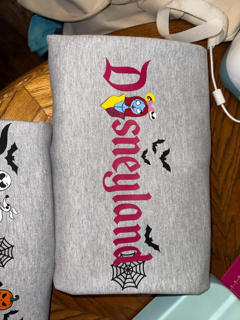 May include: Gray sweatshirt with the word "Disneyland" in a burgundy font, featuring a Sally character graphic and black bat and spiderweb designs. The sweatshirt is folded.